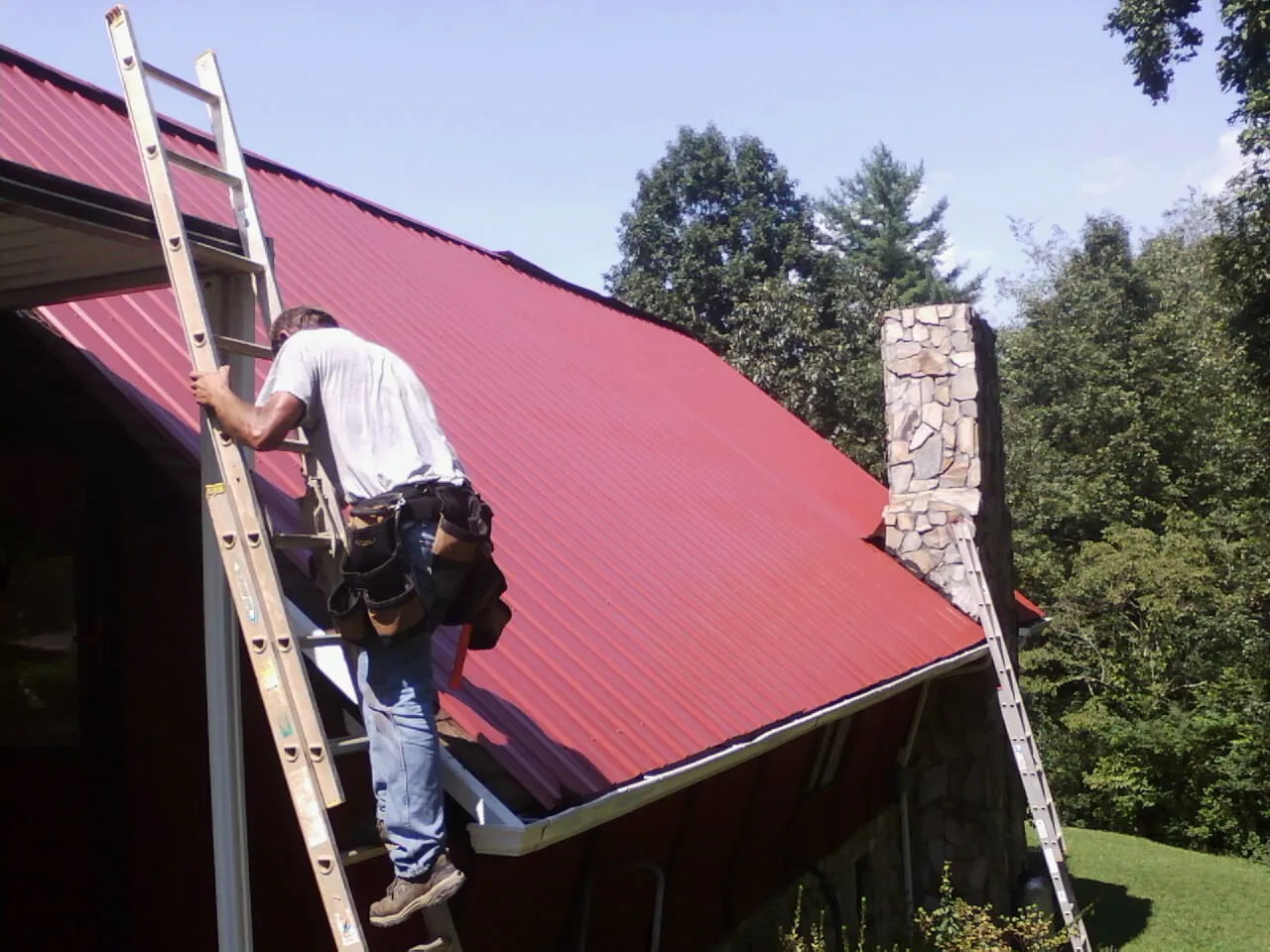 Roofer on a ladder beside a red metal roof and stone chimney — Soffit & Fascia Repair by Roofing Brothers in Morse Bluff